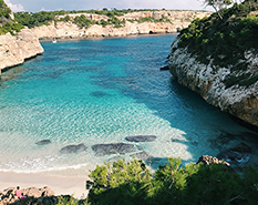 majorca beach, sandy beach at the bottow with water stretching our in front, rocky costs on all other sides with some greenery atop.