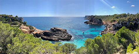 majorca cove, Sun glared blue ocean surrounded on three sides by moss, rocks, and trees, some houses on the left.